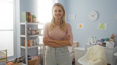 Bebek oyuncakları ve mobilyalarla çevrili, rahat bir yatak odasında kollarını kavuşturmuş gülümseyen bir kadın, neşeli bir ev ortamını resmediyor..