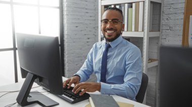 Kapalı alanda bilgisayar başında çalışan, örgülü gözlüklü, ofis ortamından emin görünen, üretkenlik ve modern iş ortamını gösteren genç bir adam.