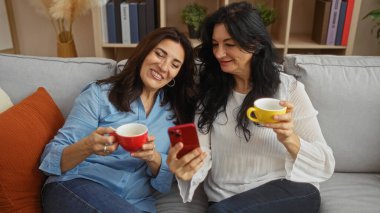 İki gülümseyen orta yaşlı kadın evlerinde rahat bir kanepede oturur, kahvelerini yukarda içerideki arkadaşlığın ve konforun özünü yakalayarak akıllı bir telefonda anlarını paylaşırlar..