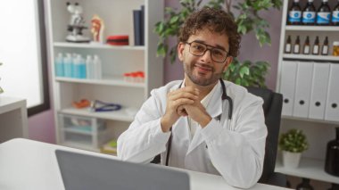 Klinikte doktor önlüğü giymiş yakışıklı genç bir adam. Arka planda tıbbi ekipmanlarla modern bir hastane odasında otururken gülümsüyor.