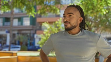 Örgü örgülü genç adam şehir parkında güneşli bir öğleden sonra etrafını çevreleyen kendinden emin bir ifade sergiliyor..