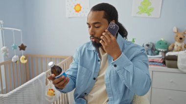 Yatak odasında sakallı genç adam bebek beşiğinin yanında ilaç şişesini incelerken telefonla konuşuyor.