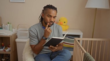 Yatak odasında oturan genç adam, bebek karyolasının yanında defter okuyor, ev ortamını yansıtıyor, yumuşak, kapalı mekan ışıklarıyla çevrili..