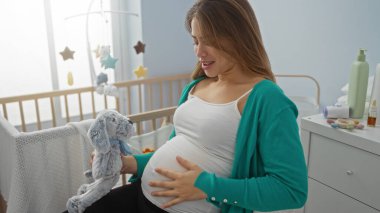 Hamile bir kadın yatak odasında oturmuş oyuncak hayvanla gülümsüyor. Etrafı bebek eşyalarıyla çevrili.