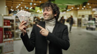 Süpermarketteki İspanyol adam Suudi riyal banknotlarına işaret ederek perakende ortamında heyecan ve finansal başarıyı vurguladı..