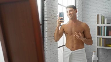 Genç İspanyol adam gülümsüyor. Aynanın önünde selfie çekiyor. Modern bir oturma odasında havluya sarılı..