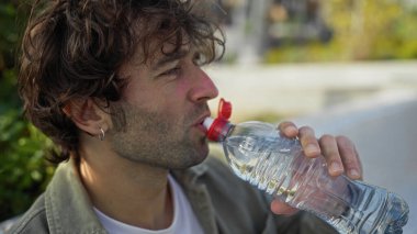 Güneşli bir günde şehir parkında su içen genç bir adam, doğa ile çevrili bir şehirde ferahlatıcı bir susuzluk anı yakalıyor..
