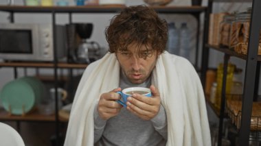 Ofiste elinde kahve dolu battaniyeyle oturan bir adam evde otururken yorgun yüz ifadelerini gösteriyor. Soğuk ya da kötü görünen mutfak eşyalarıyla çevrili.