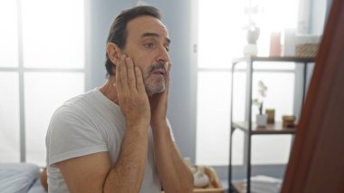 Sakin spa ortamında, hafif ışıklandırma ve minimalist dekor ile yüzünü inceleyen olgun İspanyol bir adam, sağlık ve kişisel bakım üzerine yoğunlaşmayı öneriyor.