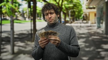 Güneşli bir şehir caddesinde İngiltere banknotlarını sayan İspanyol bir adam yeşil ağaçlar ve doğal ışık ile kentsel bir çevrede para yönetimi sergiliyor..