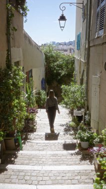 Woman walking down picturesque european alley lined with lush plants on a sunny day, embracing tranquility in an urban setting.