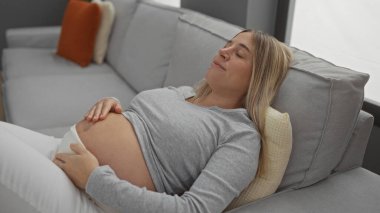 Pregnant woman relaxing on a comfortable couch in a cozy living room, gently resting her hands on her belly while expecting a baby in a peaceful home environment.