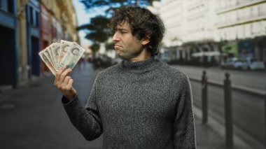 Hispanic man holding english pound banknotes on a city street outdoors, showcasing money and urban lifestyle contemplatively.