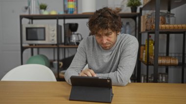 Man concentrated on tablet in a modern kitchen office setting, surrounded by shelves and kitchenware, emphasizing a relaxed workspace environment.