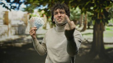 Hispanic man outdoors in a park holding polish zloty banknotes with an angry expression, emphasizing his frustration and defiance in a natural setting.