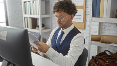 Modern bir ofiste kitap rafları ve bilgisayarlarla çevrili belgeleri okuyan genç bir İspanyol, kapalı alanda odaklanmış ve profesyonel bir atmosfer yakalıyor..