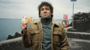 Avro banknotunu elinde tutan İspanyol bir adam deniz kıyısındaki bir plajda duruyor ve sakin sahil zemininde eşsiz bir finansal sembolizm karışımı yakalıyor..