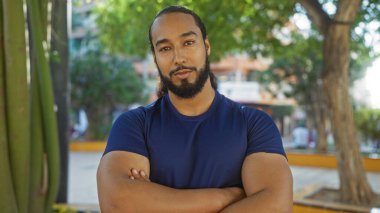 Sakallı ve çapraz kollu genç adam güpegündüz ağaçlarla ve binalarla çevrili bir şehir parkında güvenle duruyor..