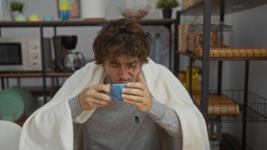 Genç adam battaniyeye sarılmış, rahat ofis mutfağında çayını yudumluyor, kendini iyi hissetmiyor ve mutfak gereçleri ve aletlerle çevrili iken sıcak bir ortam arıyor.