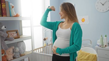 Yatak odasında hamile bir kadın, oyuncak ve kitaplarla çevrili bebek beşiğinin yanında güven içinde kolunu esnetiyor, rahat bir yuvadaki gücü ve anneliği simgeliyor..