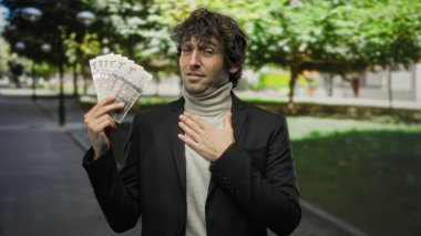 Dışarıdaki İspanyol adam bir elinde Suudi riyal banknotları tutarken diğerini göğsüne koyuyor, yemyeşil bir parkta güvenle dikiliyor..