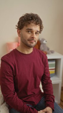 Kıvırcık saçlı, bordo tişörtlü genç adam, sıcak yatak odasında bebek beşiğinin yanında oturuyor. Düşünceli ve sakin bir ifade sergiliyor..