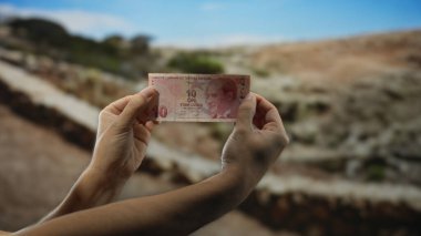 Bir park ortamında elinde 10 lira banknot tutan adam, bulanık bir bahçe arka planına karşı Türk para birimini vurguluyor..