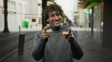 Kalabalık bir şehir caddesinde kameraya gülümseyen ve el kol hareketi yapan genç bir adam, İspanyol etnik kökenini gözler önüne seriyor ve açık şehir ortamına sahip sıradan bir moda tarzı seriyor..