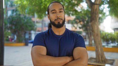 Sakallı ve sakallı bir adam şehir parkında duruyor. Kolları çaprazlanmış, ağaçlar ve şehirlerle çevrili, açık havada güç ve sakinlik yayıyor.