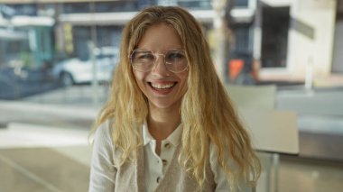 Modern ofiste gülümseyen, doğal ışık ve geniş pencereli, sarı saçlı ve gözlüklü bir kadın profesyonel ve dostane bir çalışma ortamı sergiliyor..