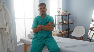 Yeşil üniformalı yakışıklı İspanyol bir adam rehabilitasyon kliniğinde kollarını kavuşturmuş, kendine güvenen bir terapi odasında poz veriyor.