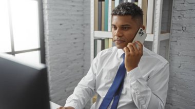 Beyaz gömlekli ve mavi kravatlı genç İspanyol bir adam çalışma odasında raflı ve pencereli akıllı bir telefonla konuşuyor.