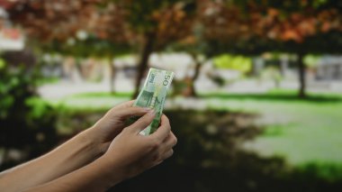 Adam bir bahçede Norveç kronu tutuyor, yeşil çevre ve doğal ışıkla çevrili bir açık hava parkında finansal işleme anını yakalıyor..