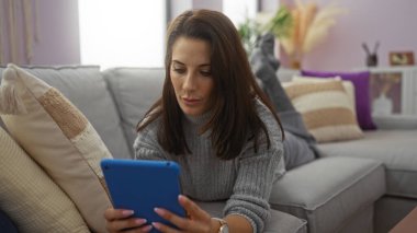 Konforlu oturma odasında rahat bir şekilde oturup tablet okuyan, modern bir dairede esmer bir kadın sergileyen şık dekorlu ve rahat bir ortam sunan bir kadın..