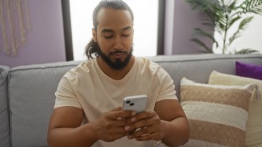 Sakallı genç adam rahat oturma odasının içinde akıllı telefon kullanıyor, modern dekorasyon ve nötr tonları var, rahat koltukta dinleniyor..