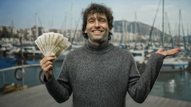 İspanyol bir adam açık deniz limanında gülümsüyor, elinde Rus rublesi var, arka planda tekneler bulanık, finans ve seyahat karışımının altını çiziyor..