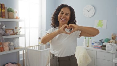 Sıcak yatak odasında kalp şeklinde jest yapan bir kadın, raflarda doldurulmuş beşiklerle, ev ortamında sıcacık gülümseyerek, sıcaklığı ve şefkati gösteriyor..