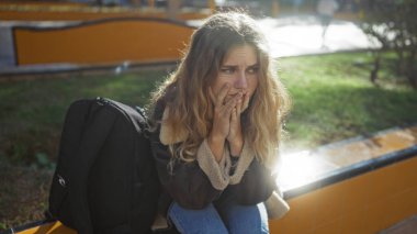 Güneşli bir günde parktaki sırt çantasının yanında elleri açık oturan kadın, duyguları ve düşüncelere dalmayı yansıtıyor..