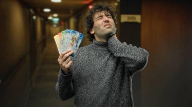 Man inside hotel reception holding swiss francs while touching sore neck, depicting discomfort in a financial setting indoors. clipart