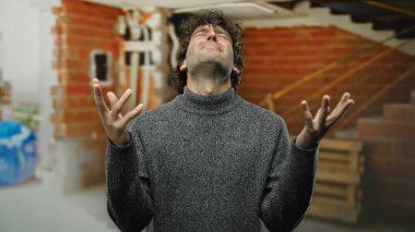 Duvarları açık, elleri kaldırmış, gri bir kazak giyen, stresi ifade eden kapalı bir inşaat alanında hayal kırıklığına uğramış genç bir adam..