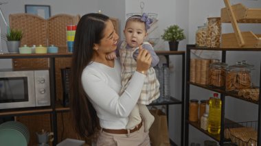 Bebek kızını yemek malzemeleriyle çevrili, rahat bir evde tutan bir kadın, modern bir ev ortamında sevgi dolu bir anne-çocuk bağı sergiliyor..