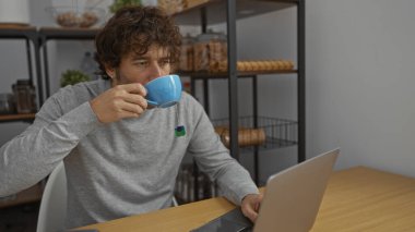 Modern ofis ortamında içki içen genç adam, düşünceli bir ifadeyle laptop ekranına odaklanmış, çağdaş dekor ile çevrili, odaklanmış çalışma ortamı öneriyor.