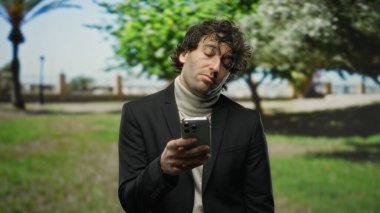 Parkta yakışıklı İspanyol bir adam telefonuna bakıyor. Yeşil ağaçların arasında üzgün ve düşünceli görünüyor..