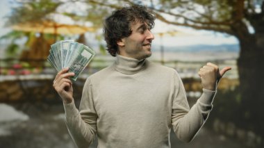 Kentin dışındaki İspanyol adam elinde bangladeş taka banknotları tutuyordu ve başparmağıyla yan tarafı göstererek gülüyordu, arka planda ağaçlar ve caddelerle yakalanmıştı..