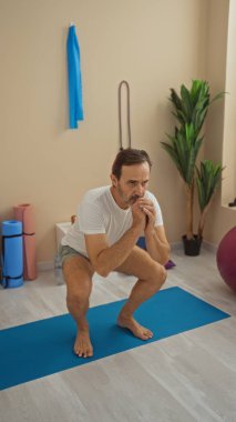 Bir sağlık merkezinde kapalı alanda egzersiz yapan bir adam, yoga minderinde çömelmiş bir şekilde duran olgun bir erkeği sergiliyor, sakin ve sakin bir ortamda spor ekipmanlarıyla çevrili..