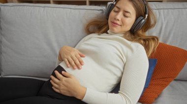 Rahat oturma odasındaki rahat koltukta kulaklıkla rahatlayan, huzur içinde dinleyen, yastığa uzanan, günlük giysiler giyen bir kadın.