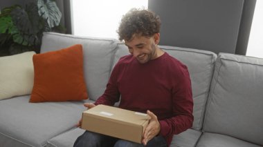 Oturma odasındaki koltukta mutlu bir şekilde oturan bir adam arka planda bitki ve yastıkların olduğu bir paket tutuyor ve bir anlık heyecanı içeride yakalıyor..