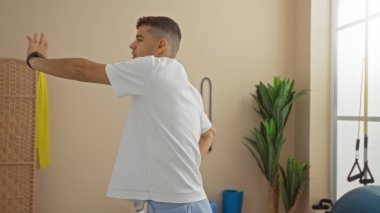 Spor salonunda egzersiz yapan genç bir adam spor aletleri ve bitkilerle odaklanmış bir duruş sergiliyor. Modern bir fitness rutinini özetliyor..