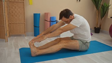 Olgun bir adam, bitkilerle ve ahşap dekorla dolu bir kaplıca odasında yoga minderinin üzerinde geriniyor. İçerideki rahatlık ve rahatlığı somutlaştırıyor..