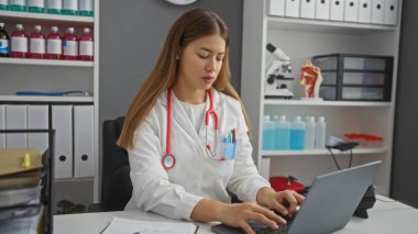Klinikte dizüstü bilgisayarı tıbbi ekipmanlarla kullanan, beyaz önlük ve steteskop giyen kadın doktor, sağlık hizmetlerinde çalışan hastaların veri girişlerine odaklandı..